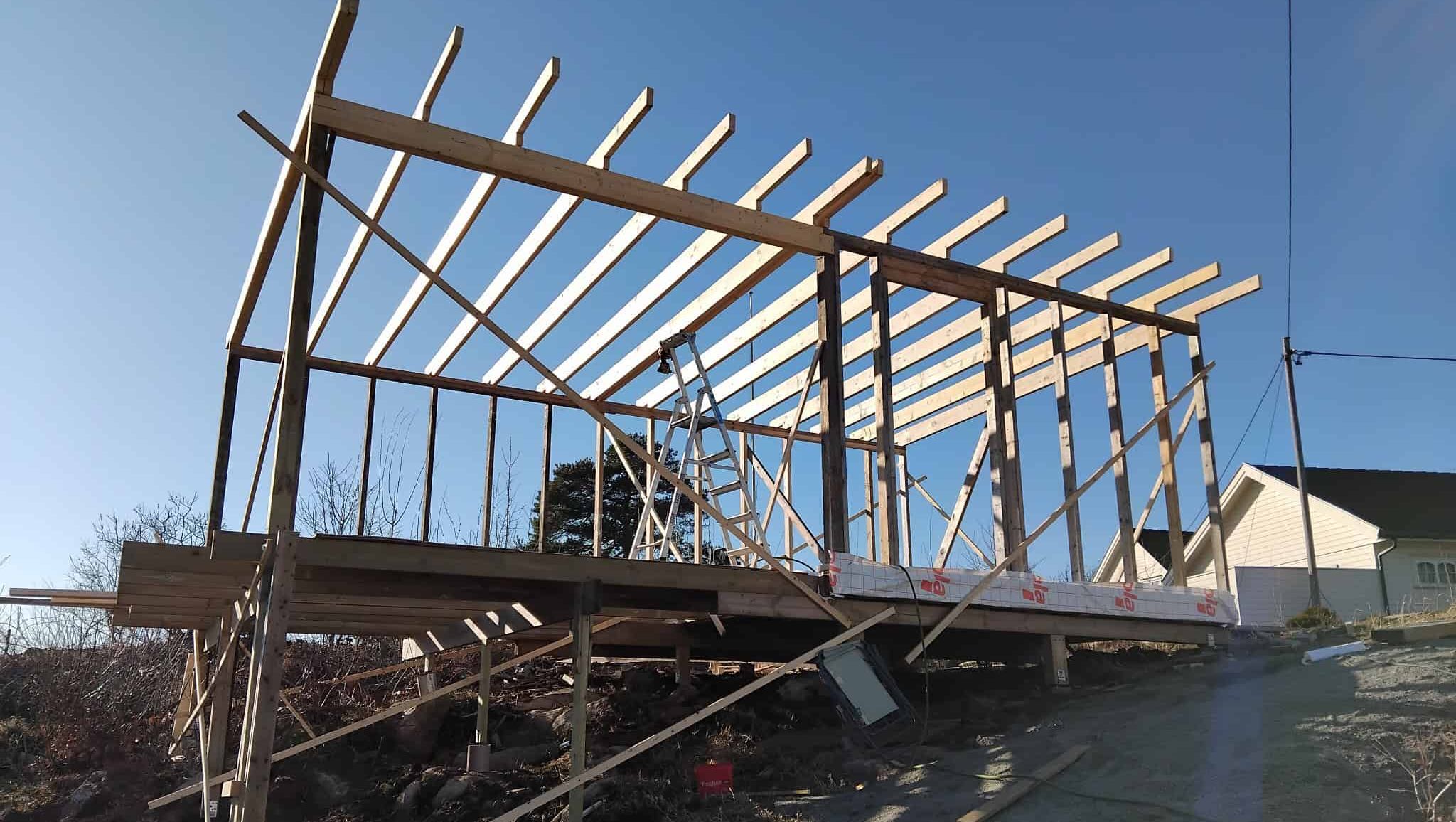 Bygning under oppføring med reisverk og takbjelker, plassert på skråtomt med støttestag og stillas.