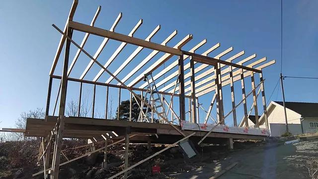 Bygning under oppføring med reisverk og takbjelker, plassert på skråtomt med støttestag og stillas.