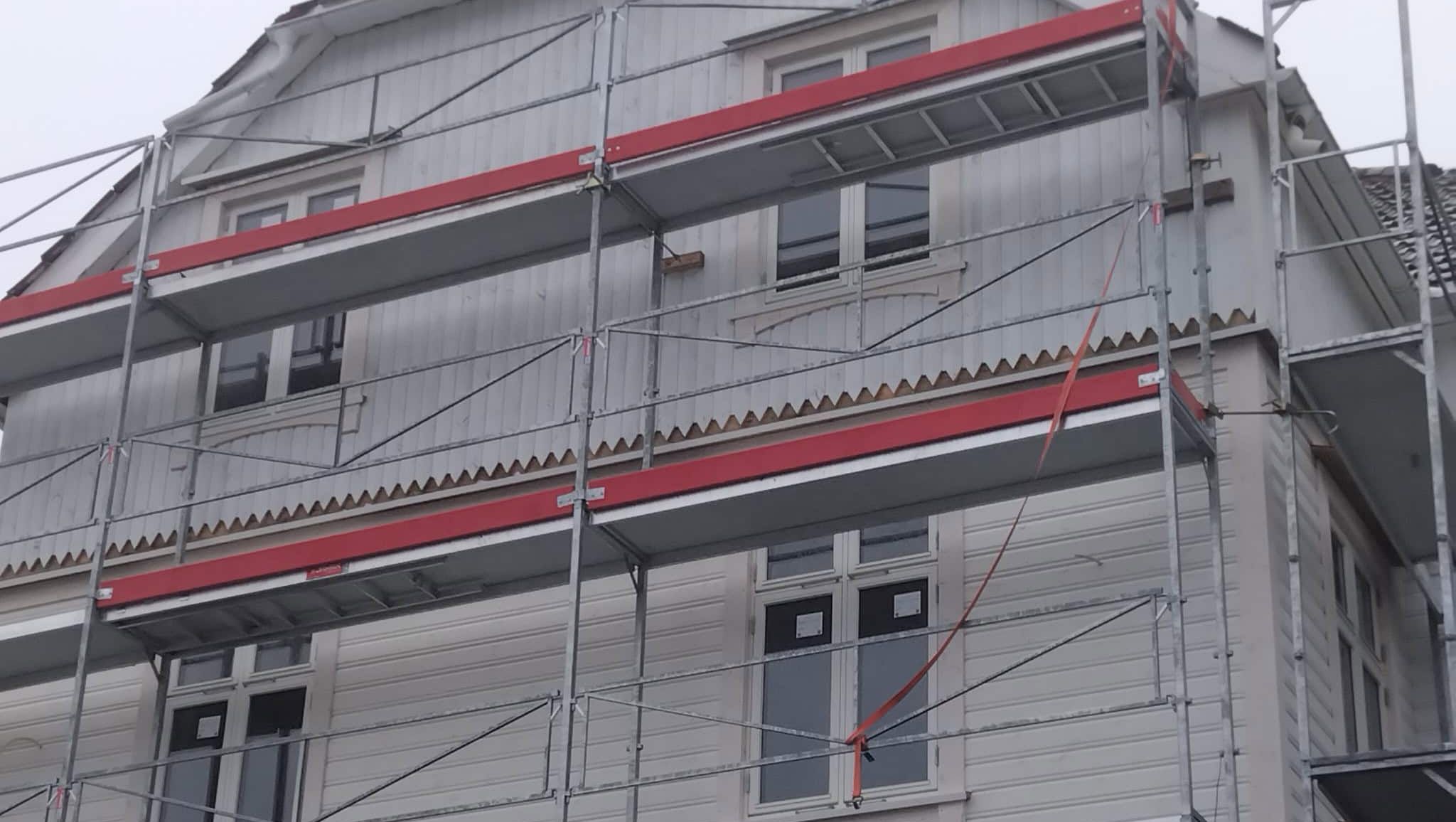 Hvit trebygning med stillas, viser fasadearbeid og oppussingsprosess.